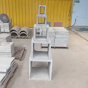 caixas de passagem em concreto, empilhadas sobre um chão de terra batida, dentro de um galpão, com outros produtos de concretos ao fundo